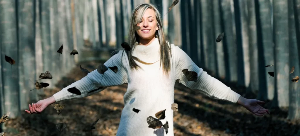 Loslassen für den Neuanfang Eine Frau in einem weißen Pullover steht in einem Wald, während Blätter um sie herum fallen. Sie breitet ihre Arme aus und lächelt.