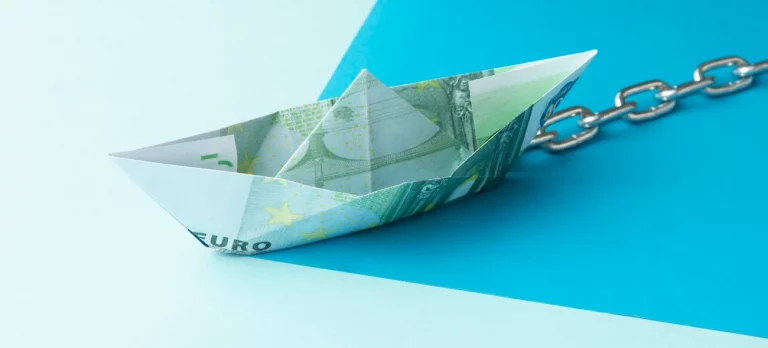Ein Papierschiff aus einem Euroschein, das mit einer Kette verbunden ist, symbolisiert die Balance zwischen Geld ausgeben und sparen.