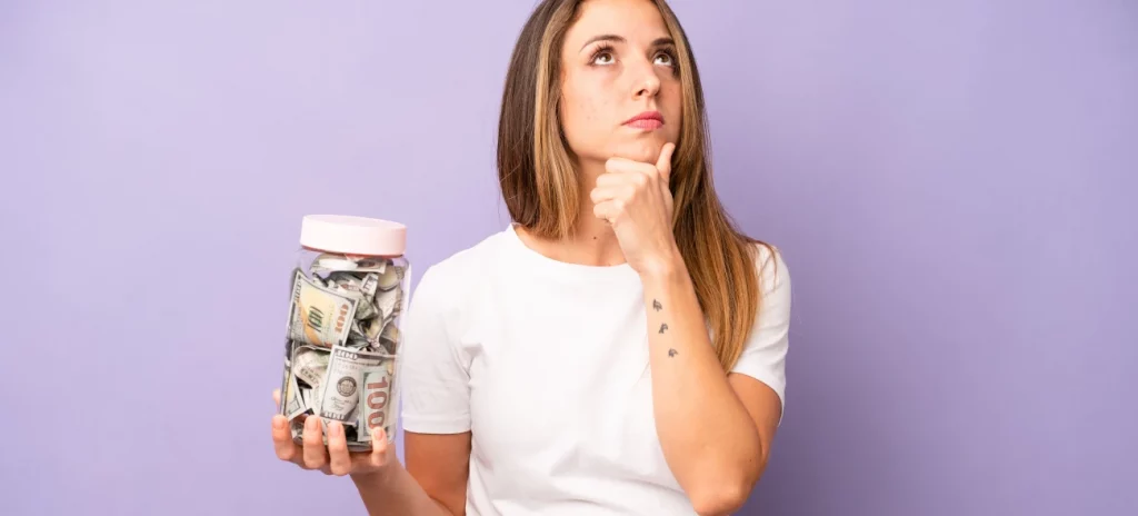 Frau denk über Geldanlagen nach und hält ein Glas voller Geldscheine und Münzen in der Hand.