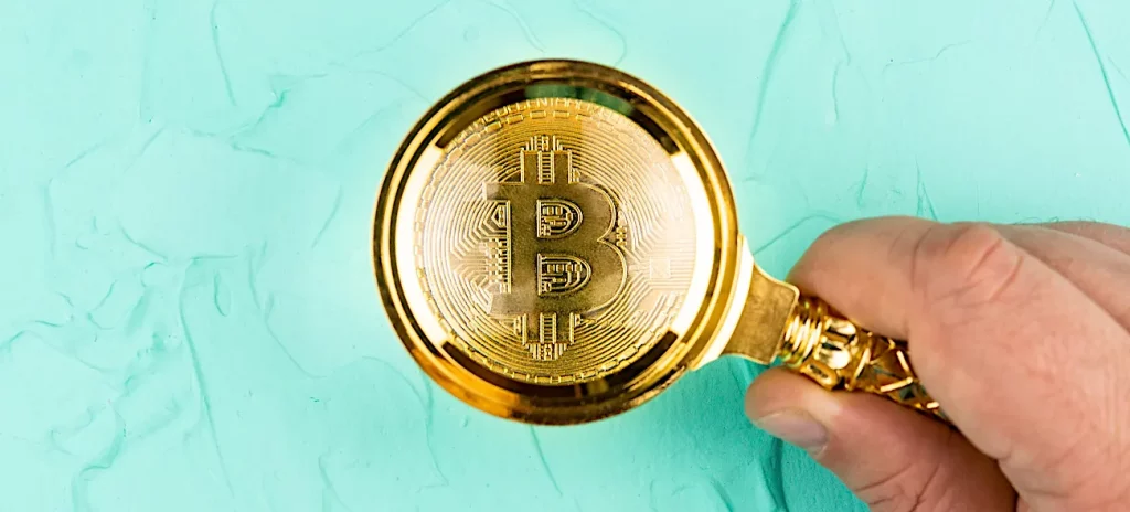 Eine Hand hält eine Lupe über eine goldene Bitcoin-Münze, die auf einem türkisfarbenen Hintergrund liegt.