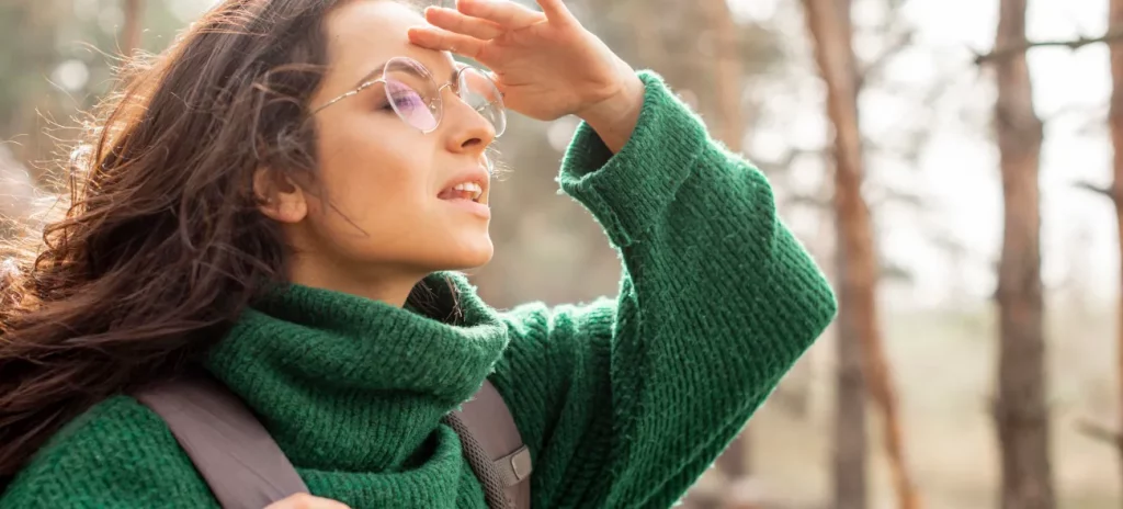 Eine junge Frau mit Brille und grünem Wollpullover ist auf der Suche nach dem Sinn des Lebens und blickt aufmerksam in die Ferne und hält eine Hand über die Stirn, um die Sicht zu verbessern. Im Hintergrund ist ein Wald zu sehen.