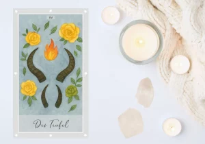 Energie der Tarotkarte „Der Teufel“. Abgebildet auf weißem Untergrund, umgeben von zwei weißen Kristallen und Teelichtern. Die Karte zeigt zwei Paar Teufelshörner, flankiert von drei gelben und einer grünen Rose sowie einer roten-orangenen Flamme in der Mitte der oberen Hörner.