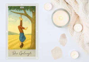 Eine Tarotkarte im Aquarellstil zeigt eine friedliche Landschaft mit einem Baum auf einem hellen Feld unter blauem Himmel. An einem Ast hängt eine Person kopfüber, der Kopf ist nicht zu sehen. Die Beine sind übereinandergeschlagen, was der Szene eine ruhige, meditative Ausstrahlung verleiht.